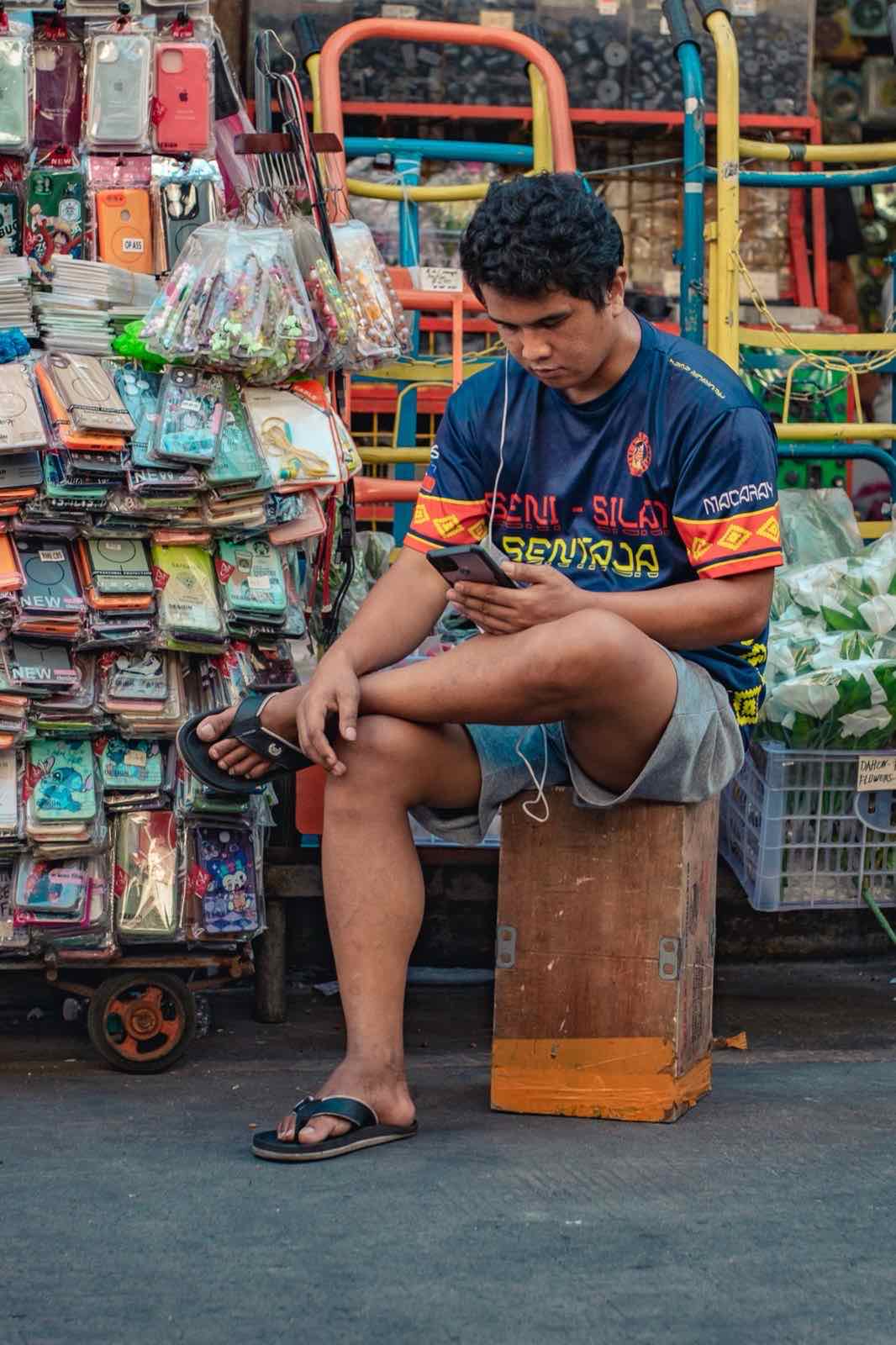 A street vendor sits among colorful merchandise checking a smartphone in Manila.