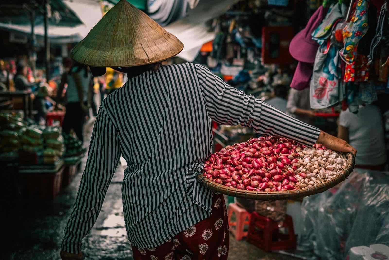 A woman carrying a basket of fresh vegetables through a produce market.